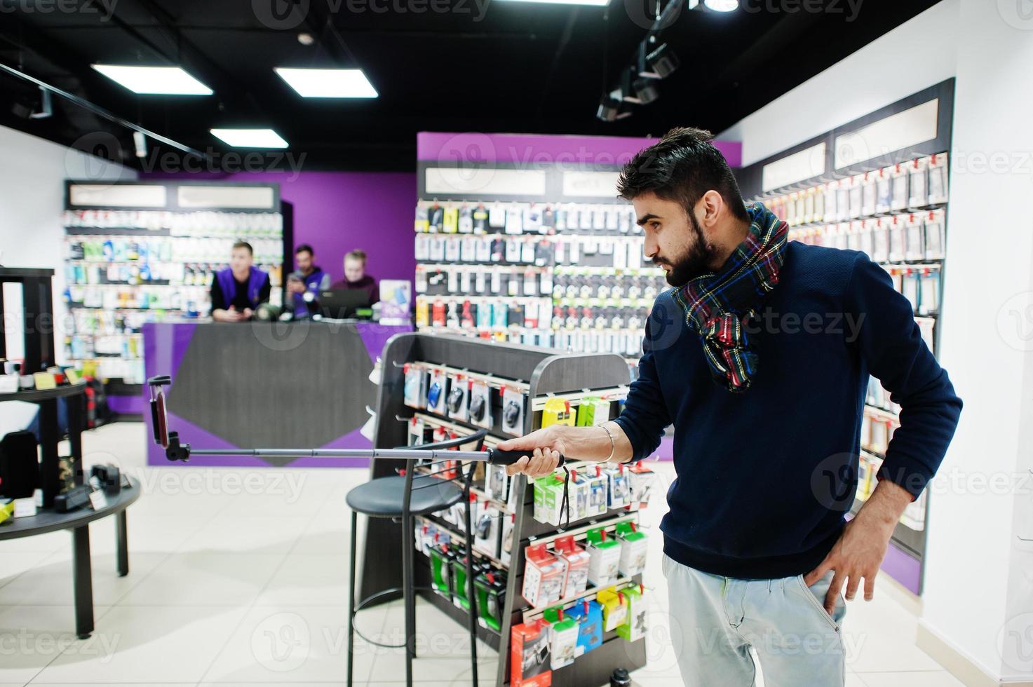 Indian man customer buyer at mobile phone store making selfie by smartphone on monopod stick. South asian peoples and technologies concept. Cellphone shop. photo
