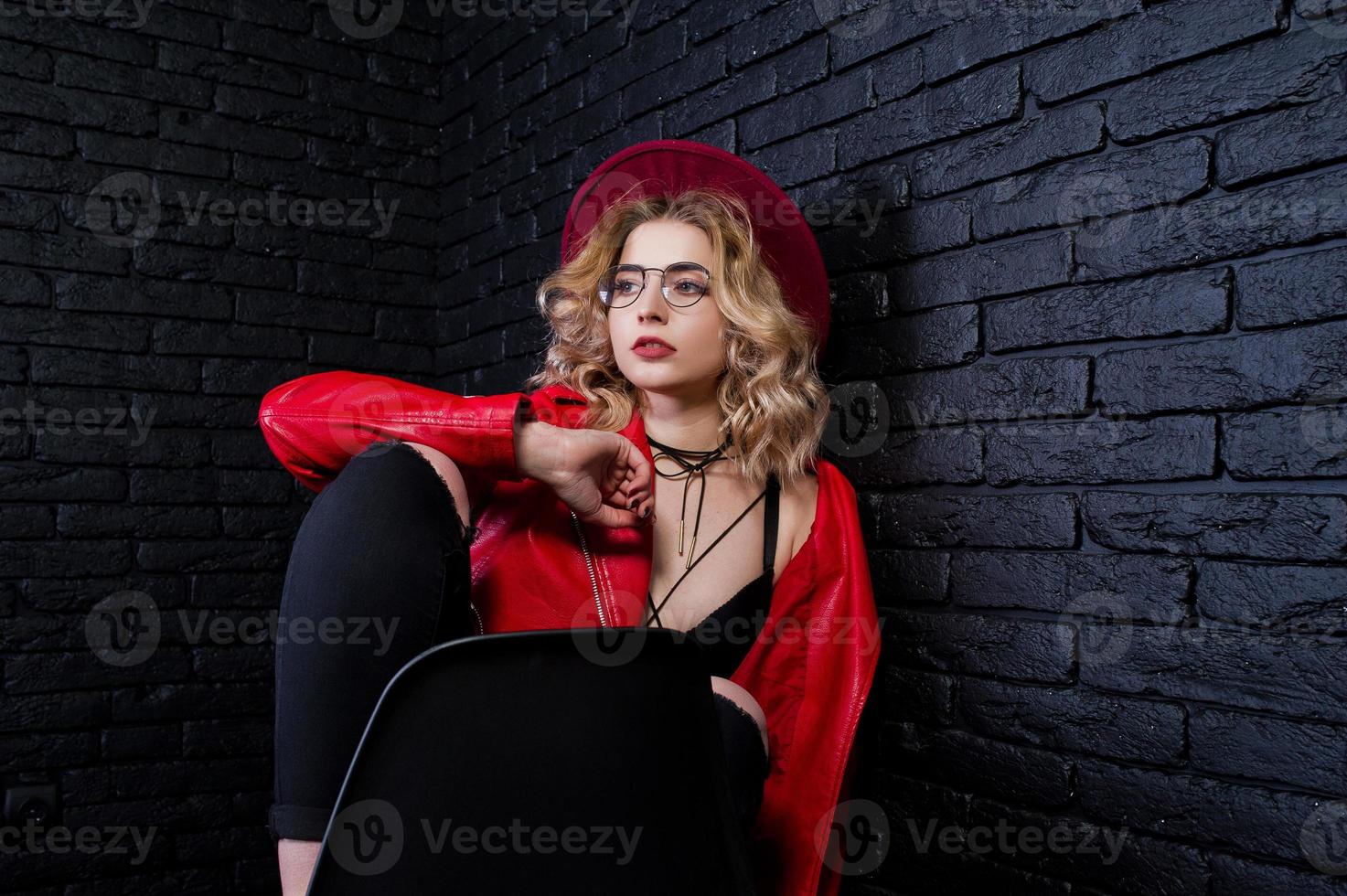 Studio portrait of blonde girl in red hat, glasses and leather jacket posed on chair against brick wall. photo