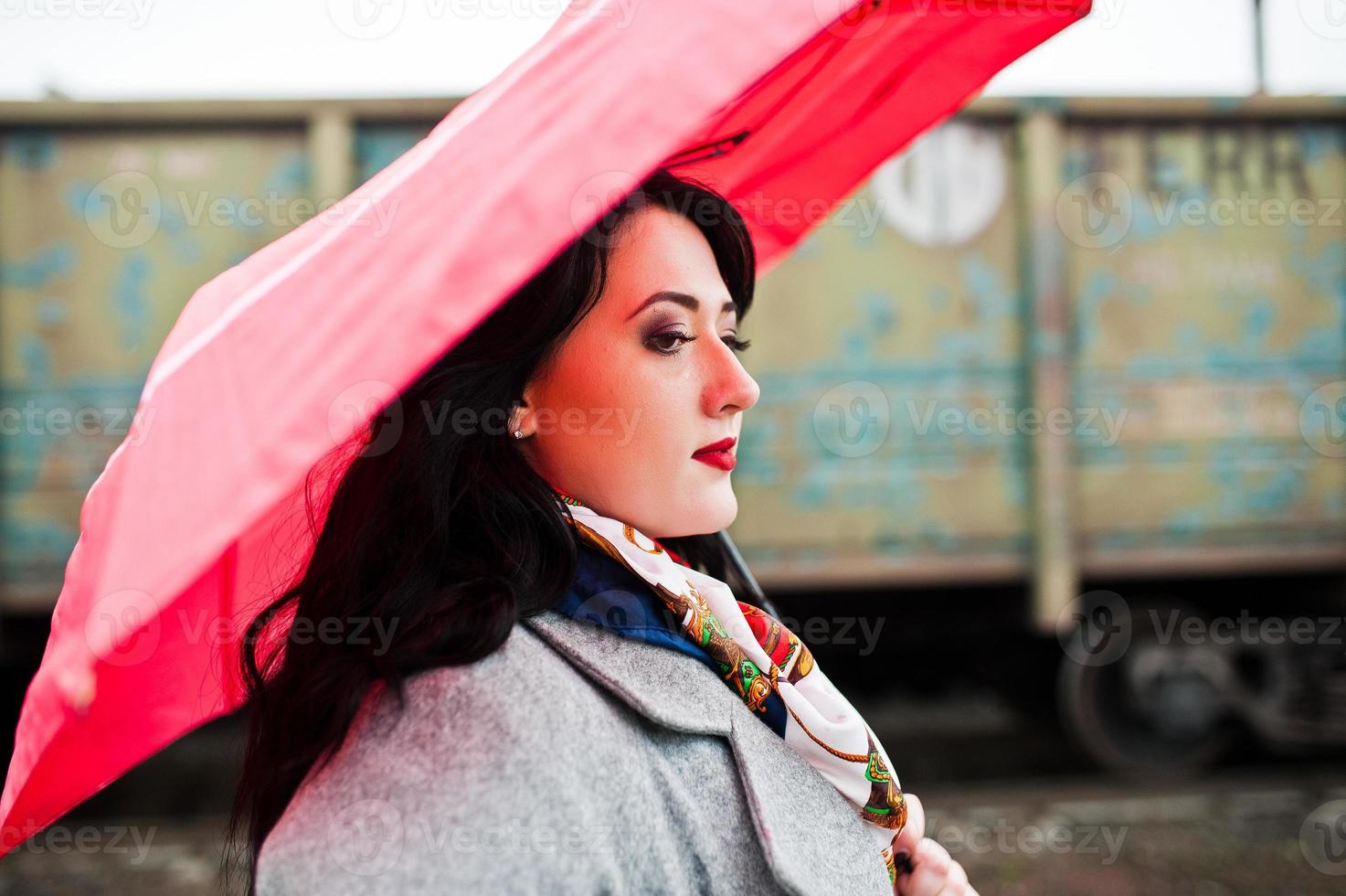 Brunette girl in gray coat with red umbrella in railway station. photo