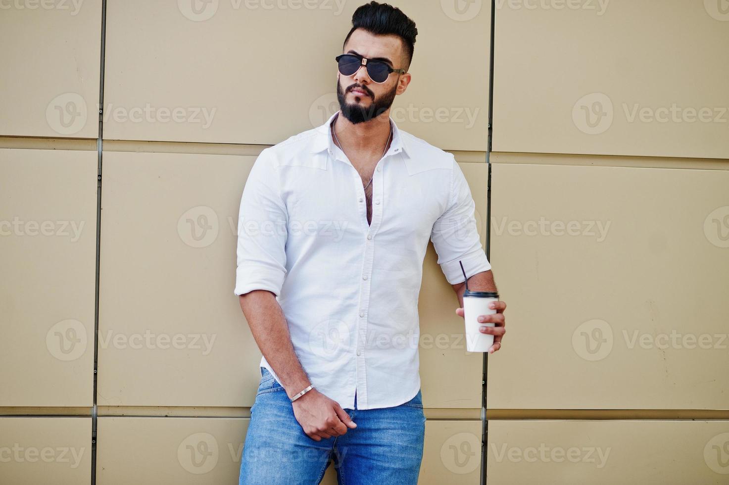 Stylish tall arabian man model in white shirt, jeans and sunglasses posed at street of city. Beard attractive arab guy with cup of coffee against golden wall. photo