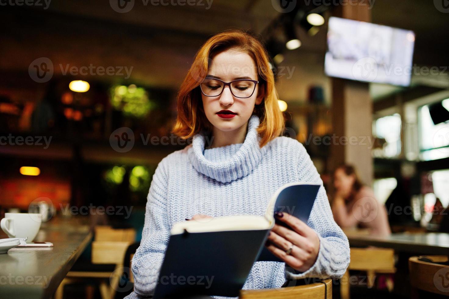 Cheerful young beautiful redhaired woman in glasses sitting at her working place on cafe reading something at her notebook. photo