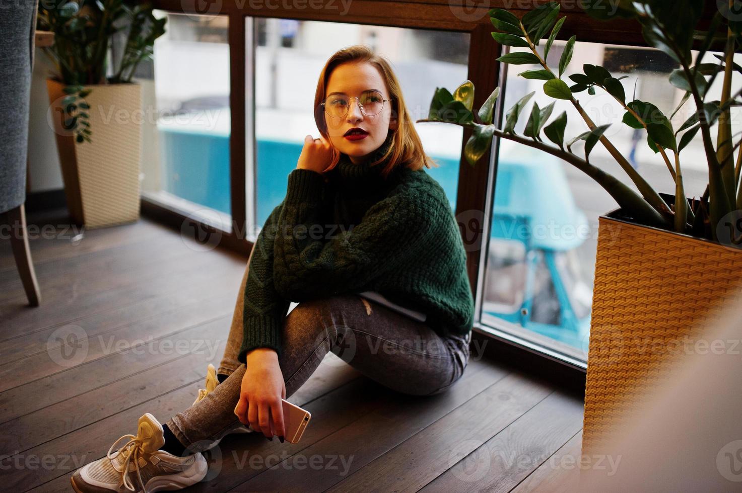 Cheerful young beautiful redhaired woman in glasses, green warm wool sweater, using her notebook, while sitting on cafe against window. She sit on floor and think about something. photo