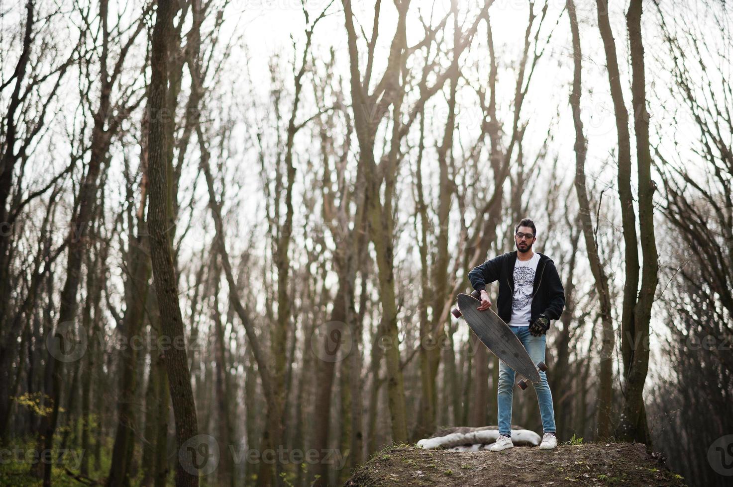 Street style arab man in eyeglasses with longboard on wood. photo