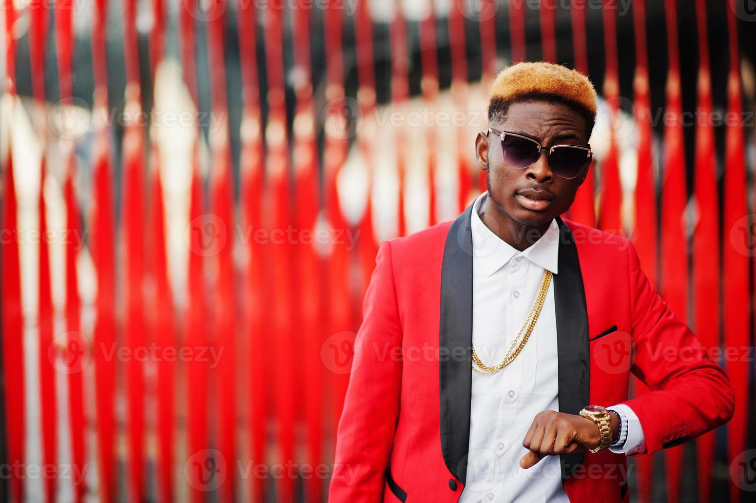Fashion african american man model at red suit, with highlights hair and handbag posed at street and looking at his watches. photo