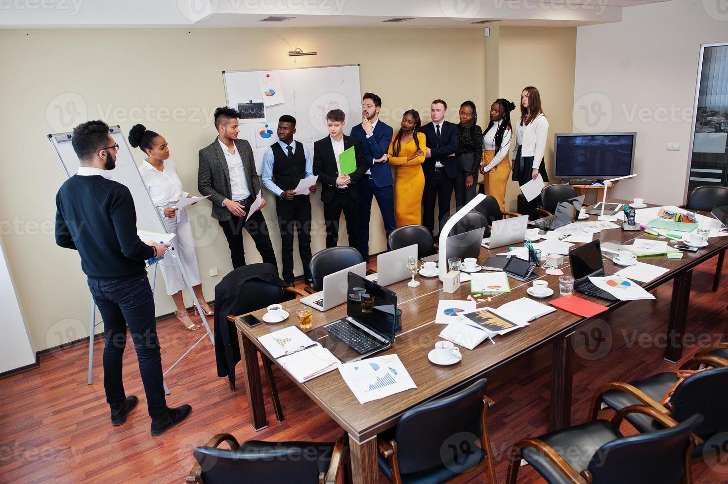 Mixed race business coach presenting report standing near whiteboard pointing on sales statistic shown on diagram and chart teach diverse company members gathered together in conference room. photo