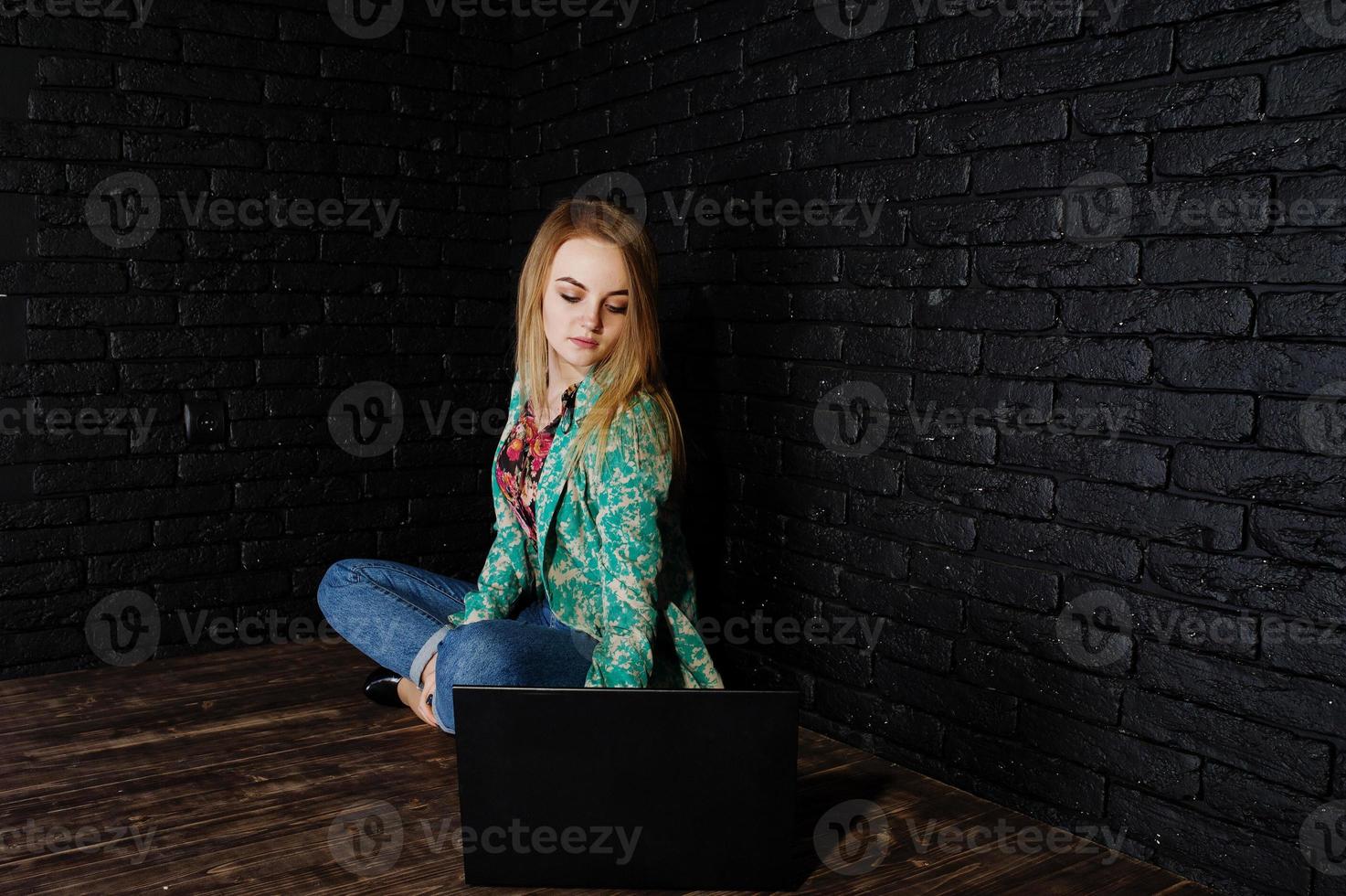 Stylish blonde girl in jacket and jeans with laptop against brick black wall at studio. photo