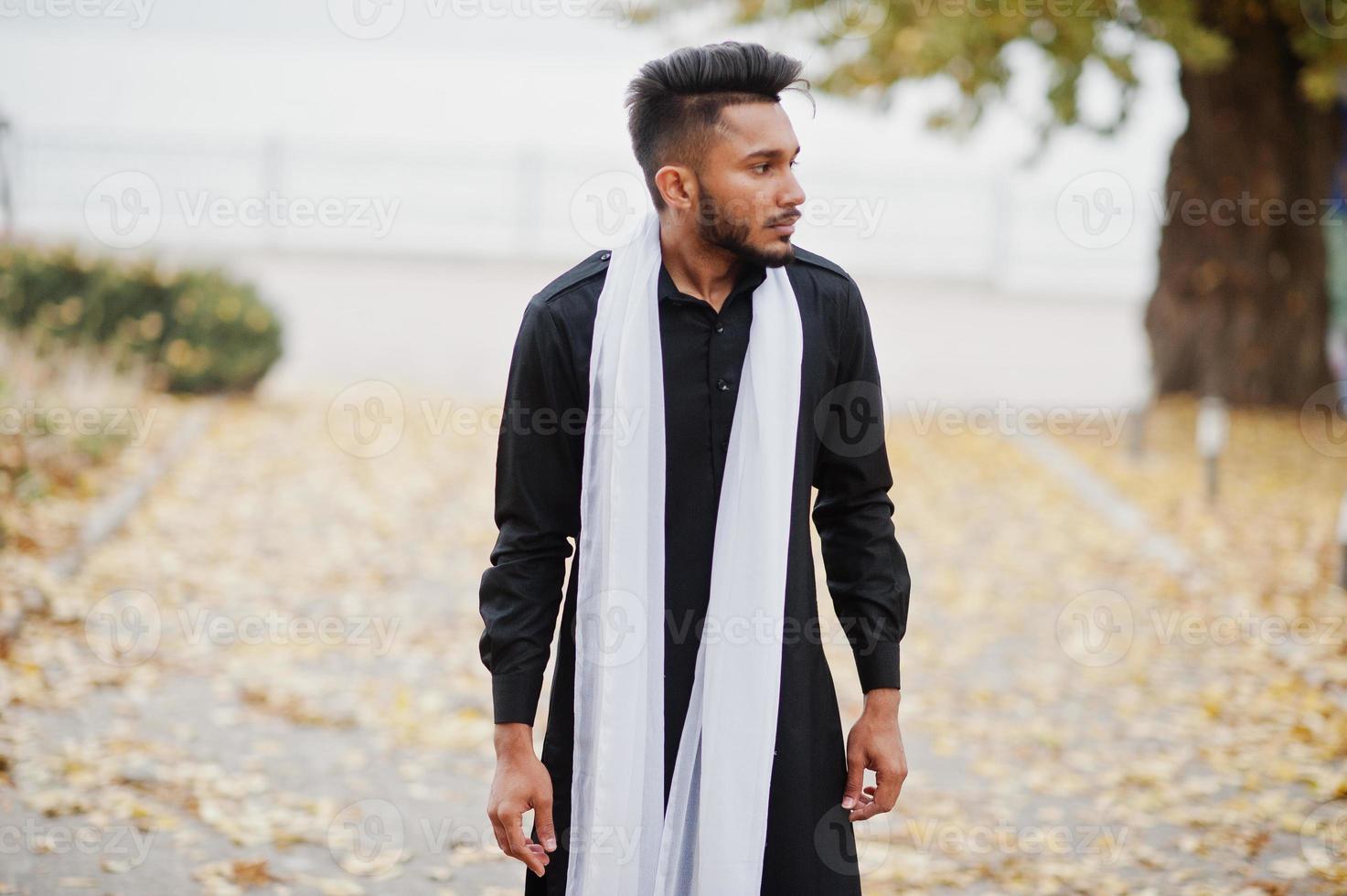 Indian stylish man in black traditional clothes with white scarf posed outdoor against autumn leaves tree. photo