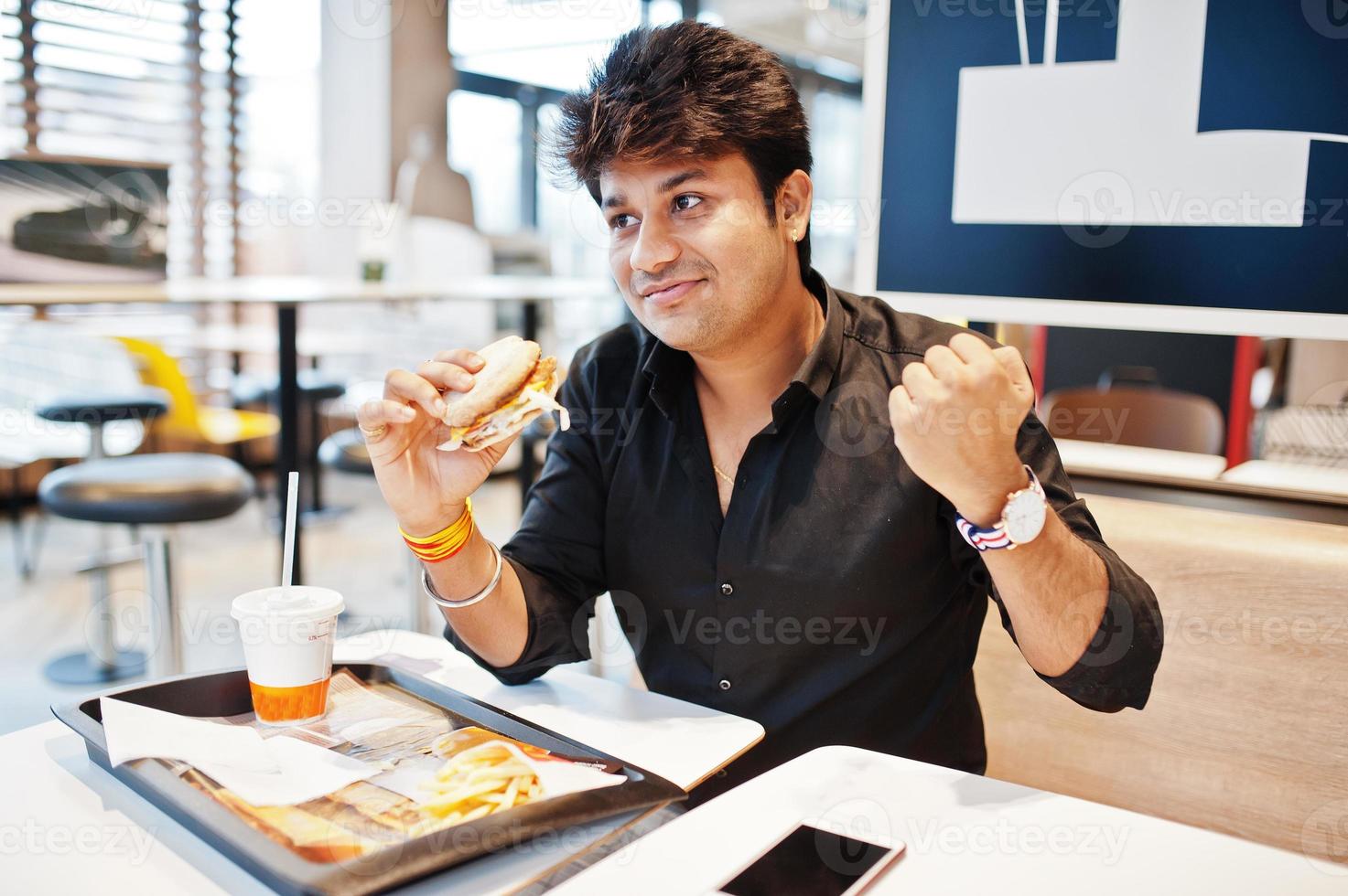 Stylish indian man sitting at fast food cafe and eating hamburger, read morning news on mobile phone. He is happy and show like him win something. photo
