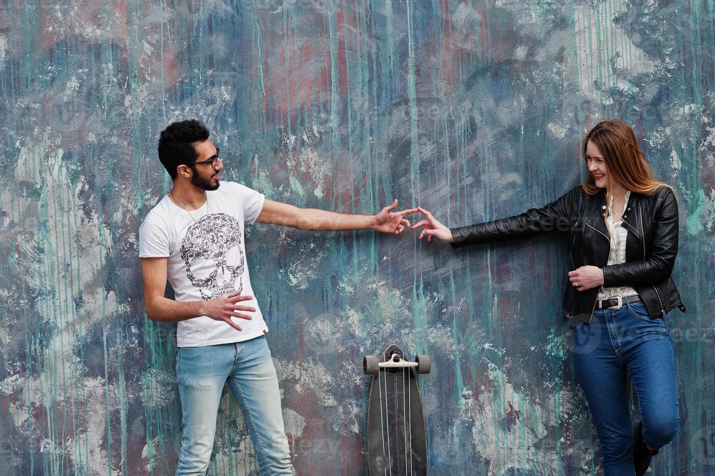 Cool multiracial couple posing against wall with longboard and touching fingers each other. photo