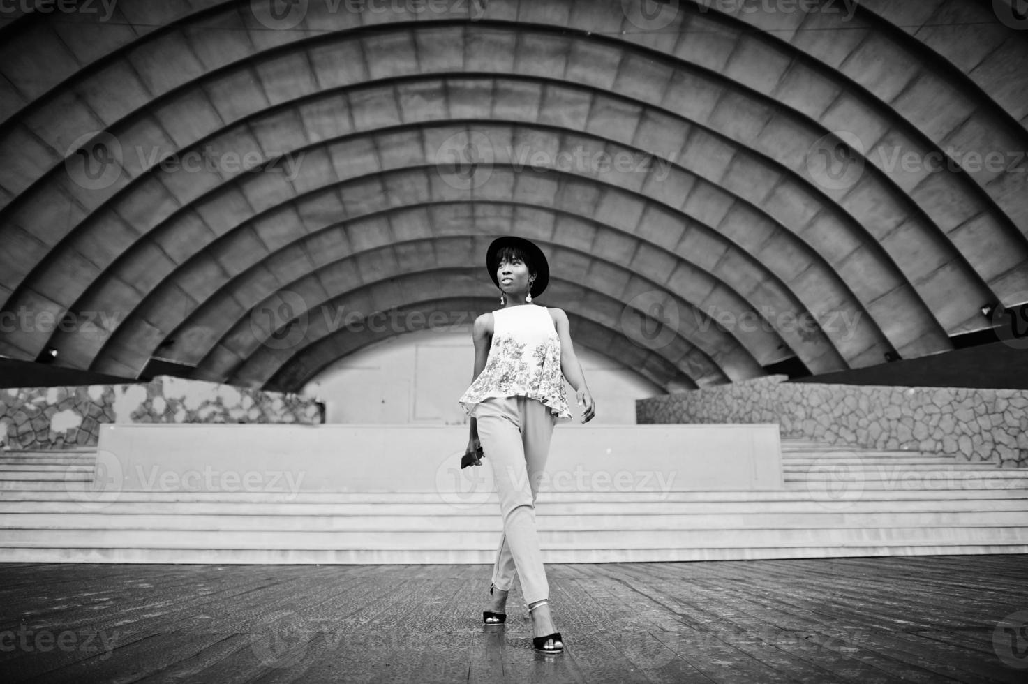 Amazing african american model woman in green pants and black hat posed outdoor against arena hall. photo