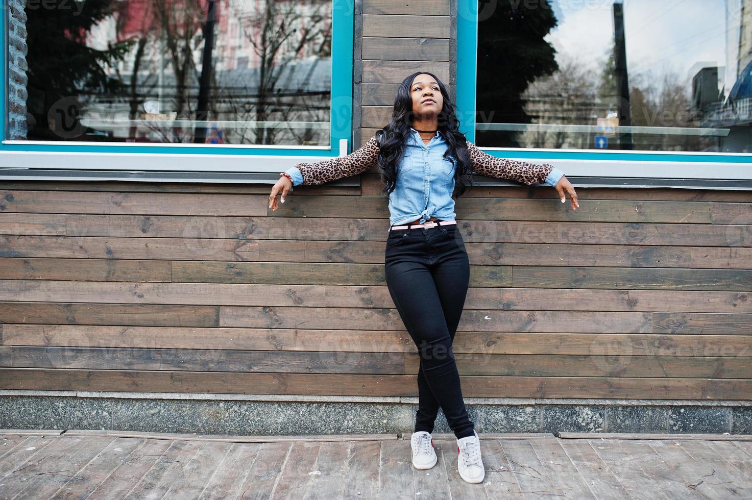 chica afroamericana hipster con camisa de jeans con mangas de leopardo posando en la calle contra una casa de madera con ventanas. foto
