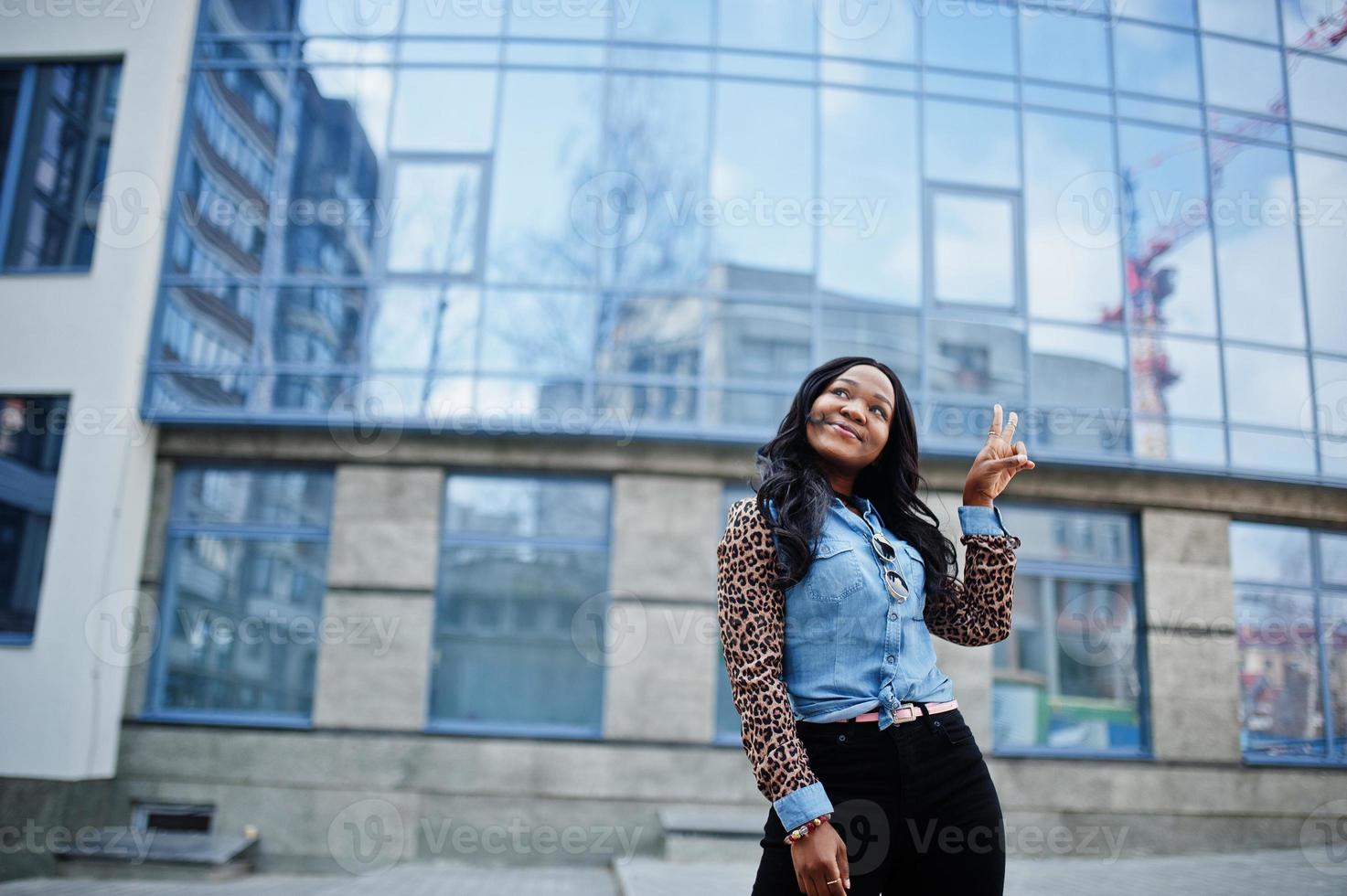 Hipster african american girl wearing jeans shirt with leopard sleeves posing at street against modern office building with blue windows. photo
