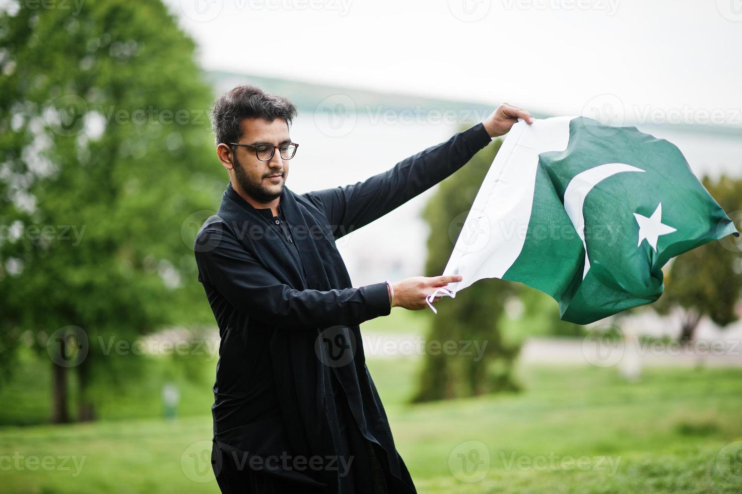 Pakistano bangladesh man wear black traditional clothes and eyeglasses pose outdoor, holding Pakistan flag. photo