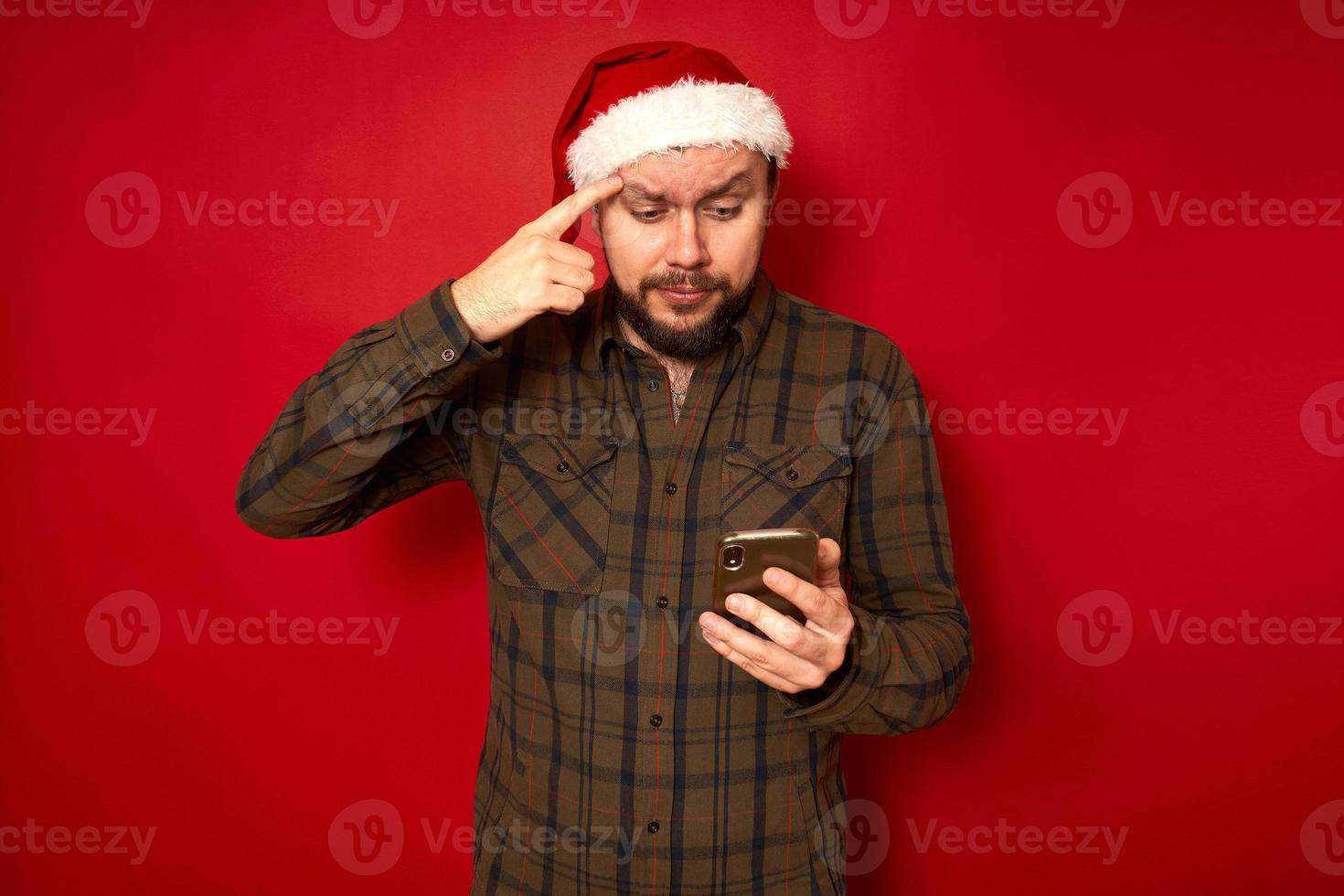 thoughtful serious man in xmas hat reads on phone holds index finger to eyebrow tries to understand conditions isolated studio background concept - holidays, technology, social networks, communication photo