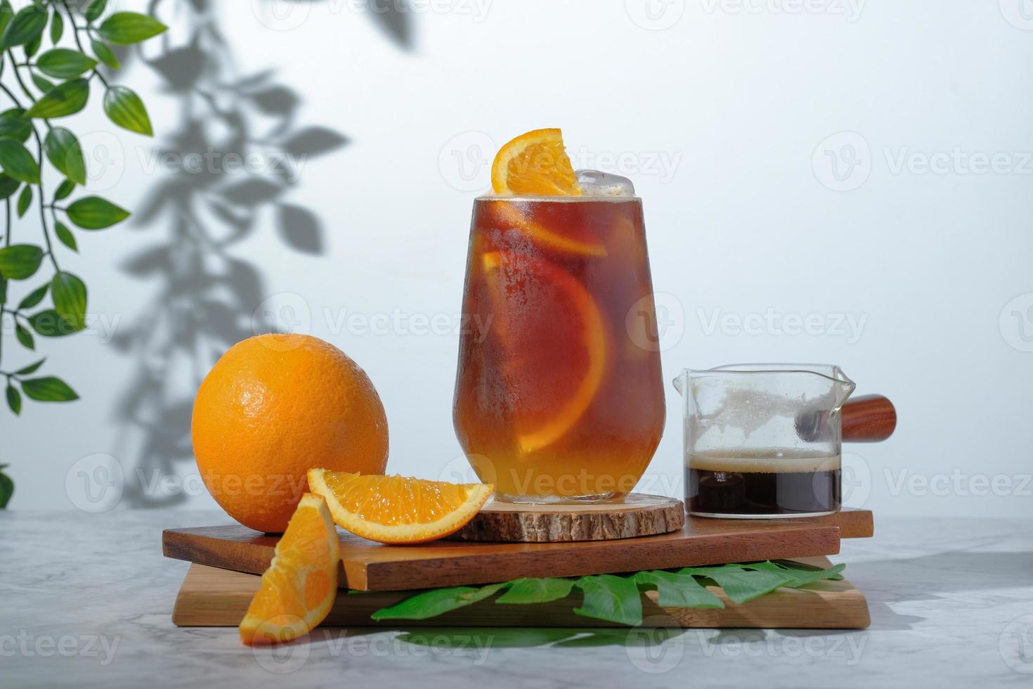 café negro helado frío mezclado con jugo de naranja y rodajas de naranja en vidrio sobre placa de madera en fondo blanco con sombra de silueta de hojas naturales. bebida fresca de verano de dos tonos por la mañana foto