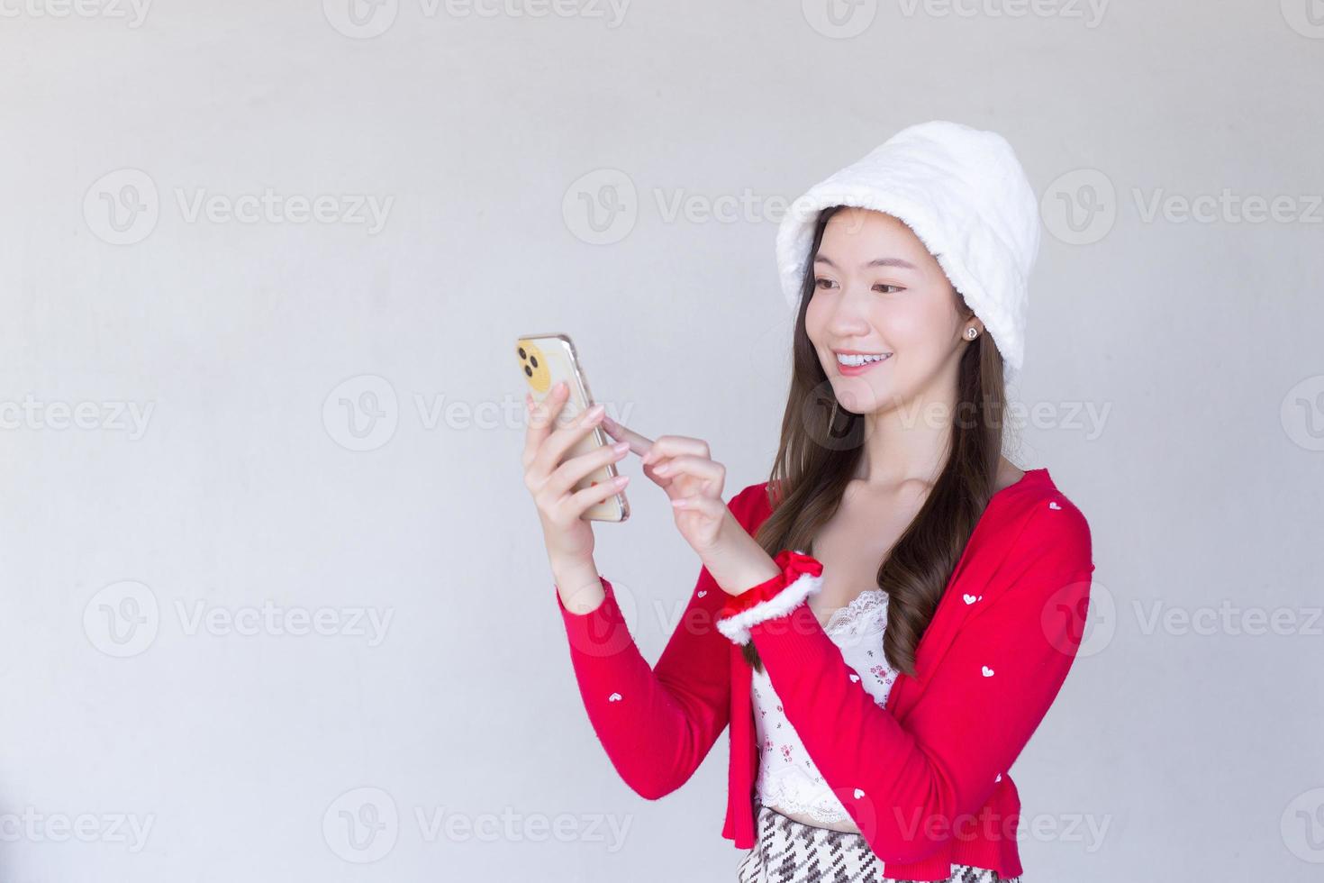 Portrait of a pretty Asian teen girl wearing a red dress and white hat happily using a smartphone on a white background. photo