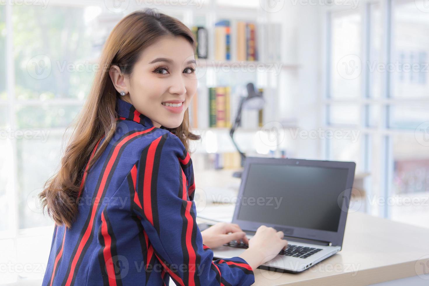 una mujer asiática confiada está descansando su mano en el teclado de la computadora portátil y voltea su rostro con una sonrisa en una sala de trabajo en casa. foto