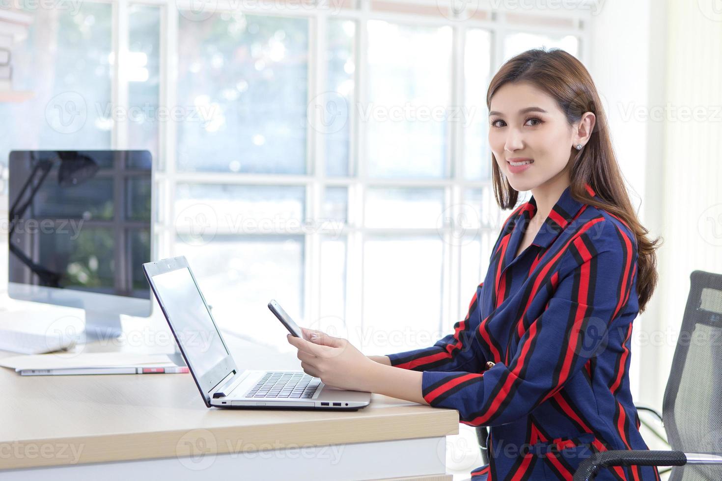 una mujer asiática sostiene el teléfono móvil en sus manos con una cara sonriente en la oficina mientras hay una computadora portátil y una computadora sobre la mesa. foto