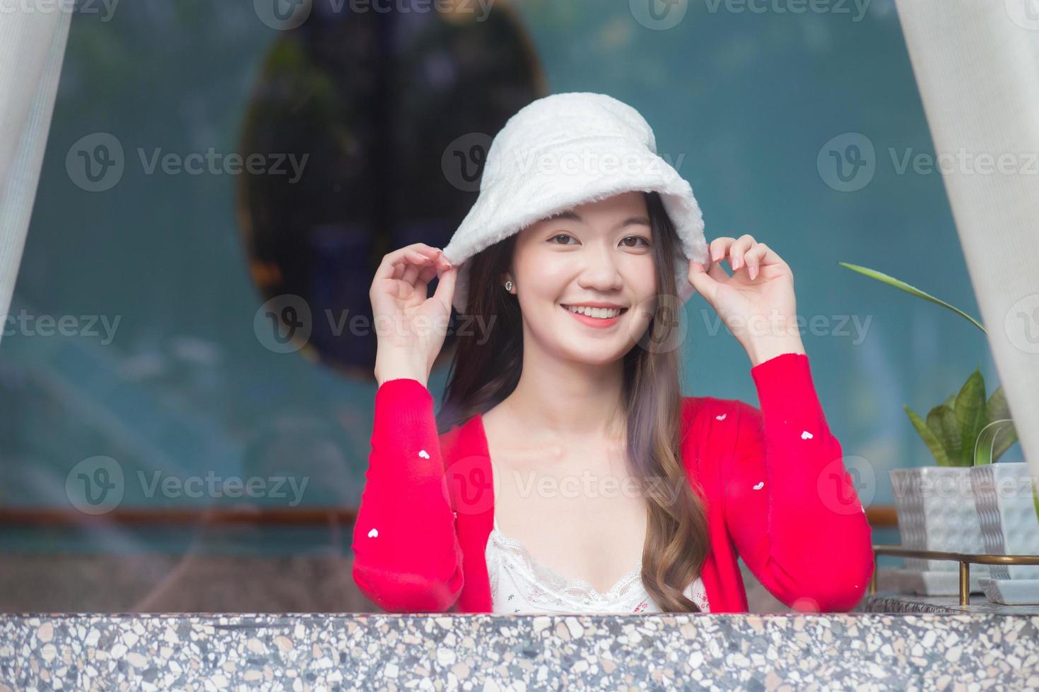 hermosa mujer asiática usa un sombrero blanco y una camisa roja mientras se sienta cerca de una ventana acristalada en el tema de año nuevo e invierno. foto