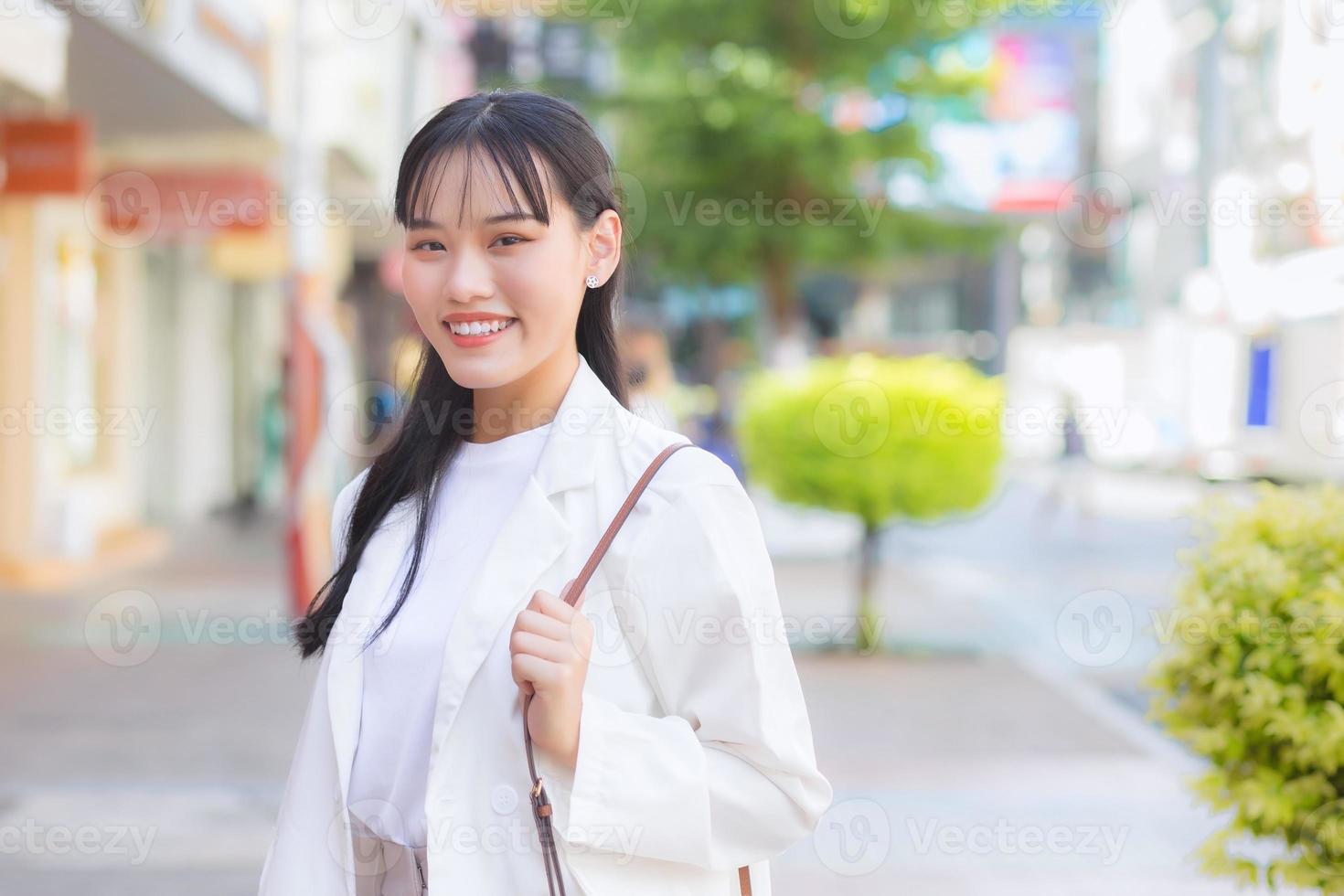 Confident young business Asian working woman who wears a white shirt and shoulder bag smiles happily while she is walking to work at office in the city. photo
