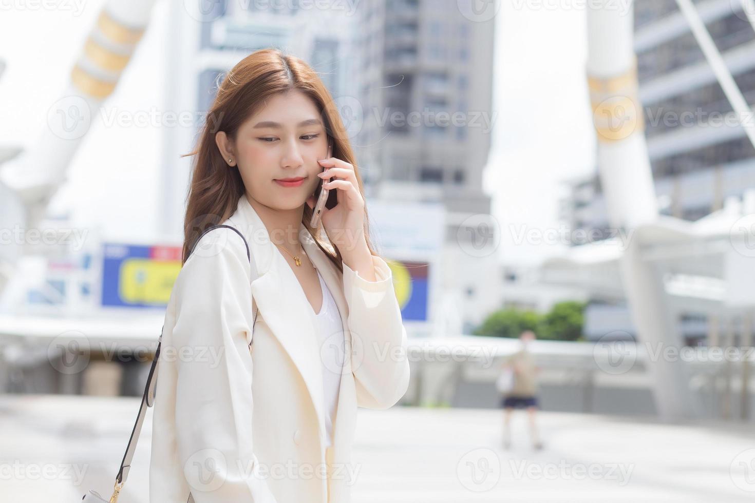 Young Asian business woman in white suit smiles and holds a smartphone in her hands in happily workday at the city as a background. photo