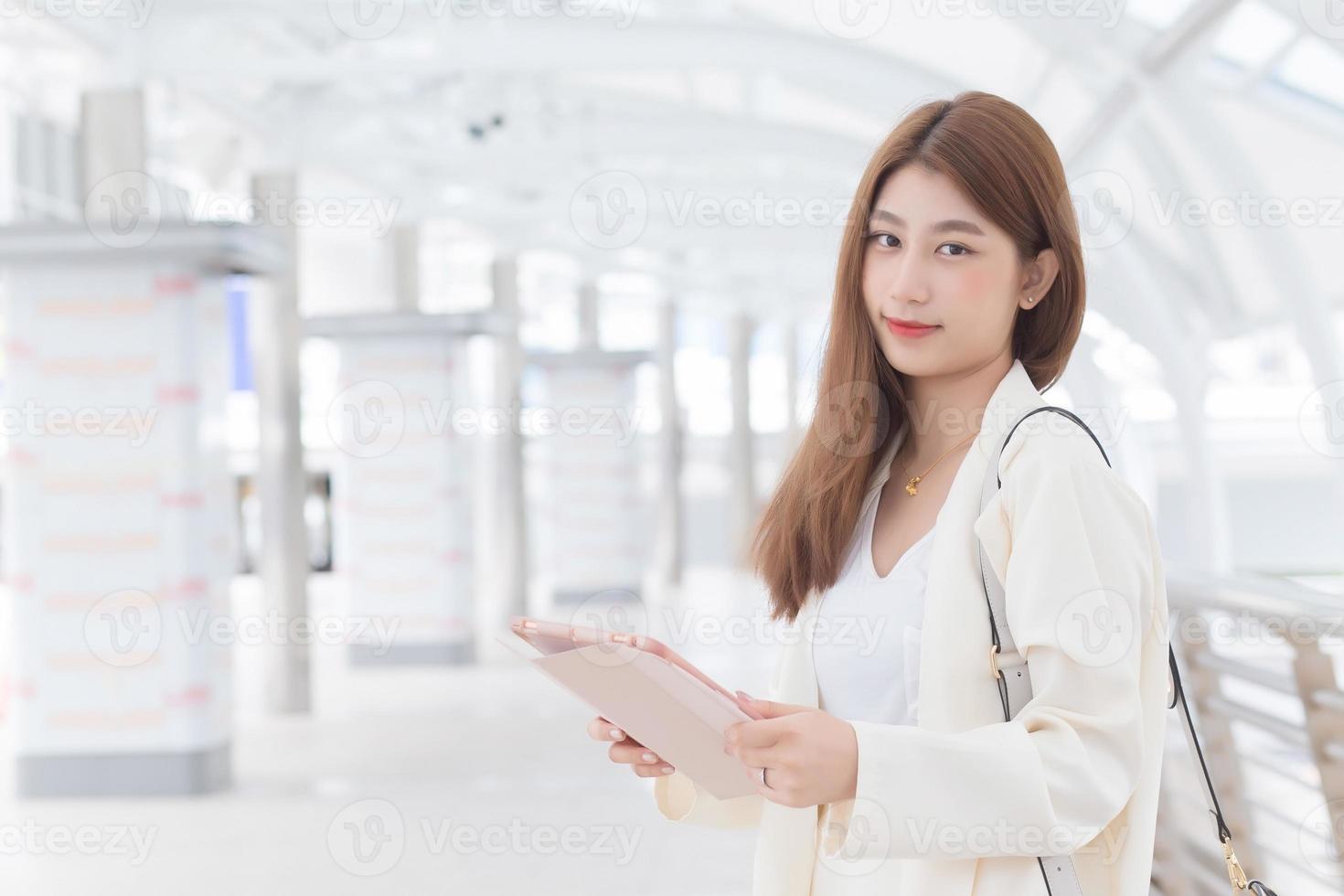 Beautiful young age Asian working business woman in white suit is outdoors to meet a client at the office her holding hand tablet to find information with a city background. photo