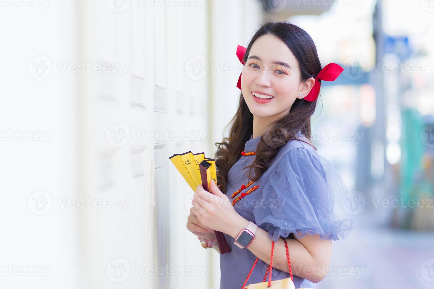 A beautiful Asian woman in a gray Qipao dress with a red bow is smiling, holding a fan on the street in Chinatown, Thailand. photo