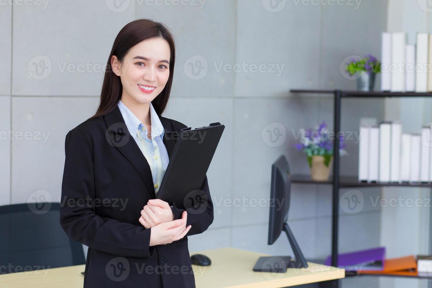 una joven empresaria asiática profesional con traje negro sonríe alegremente mientras trabaja y sostiene el portapapeles con confianza en el cargo. foto