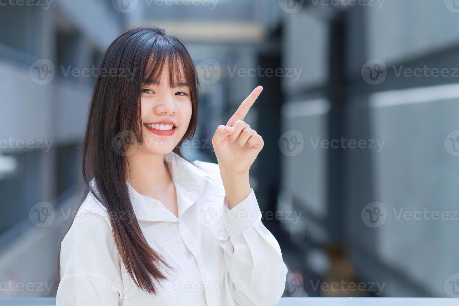 Professional young Asian working woman who wears white shirt is pointing hand to present something while outdoors in the city with an office building in the background. photo