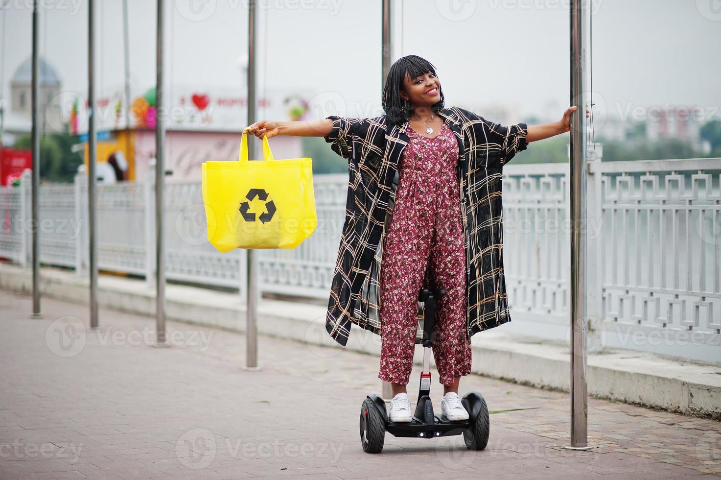 Beautiful african american woman using segway or hoverboard. Black girl with yellow  cloth eco bags recycling symbol. photo