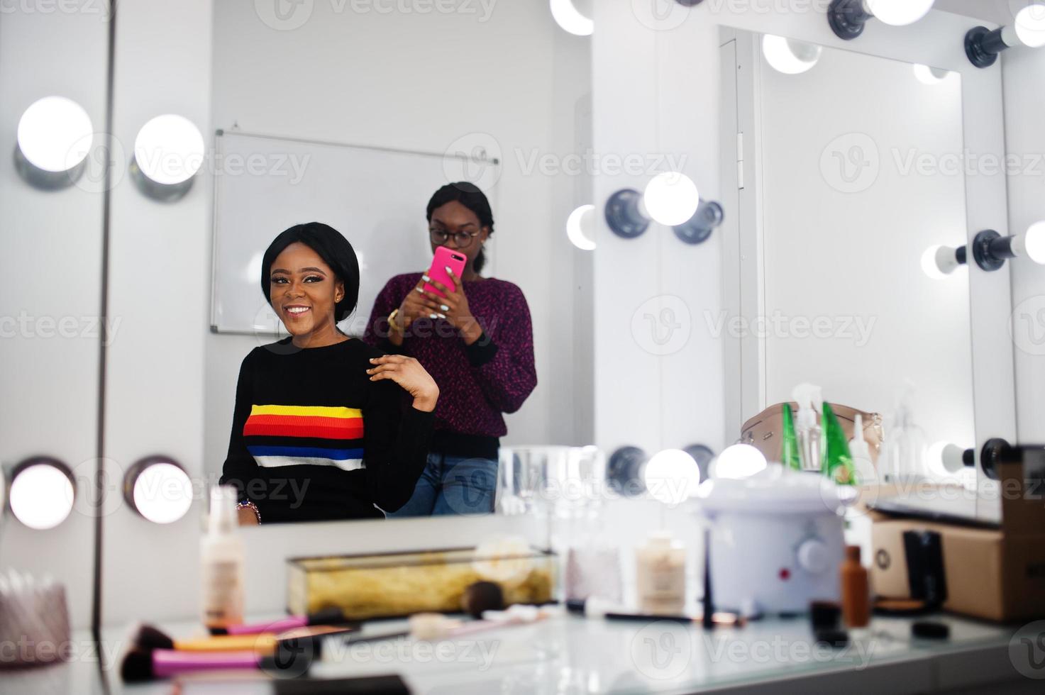 African American woman applying make-up by make-up artist at beauty saloon. Artist make photo on mobile phone of her work.