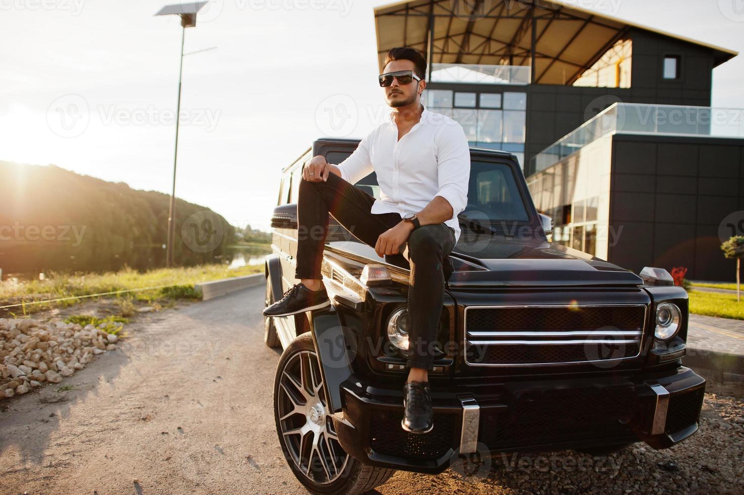 Solid asian man in white shirt and sunglasses posed near black mafia suv car. photo