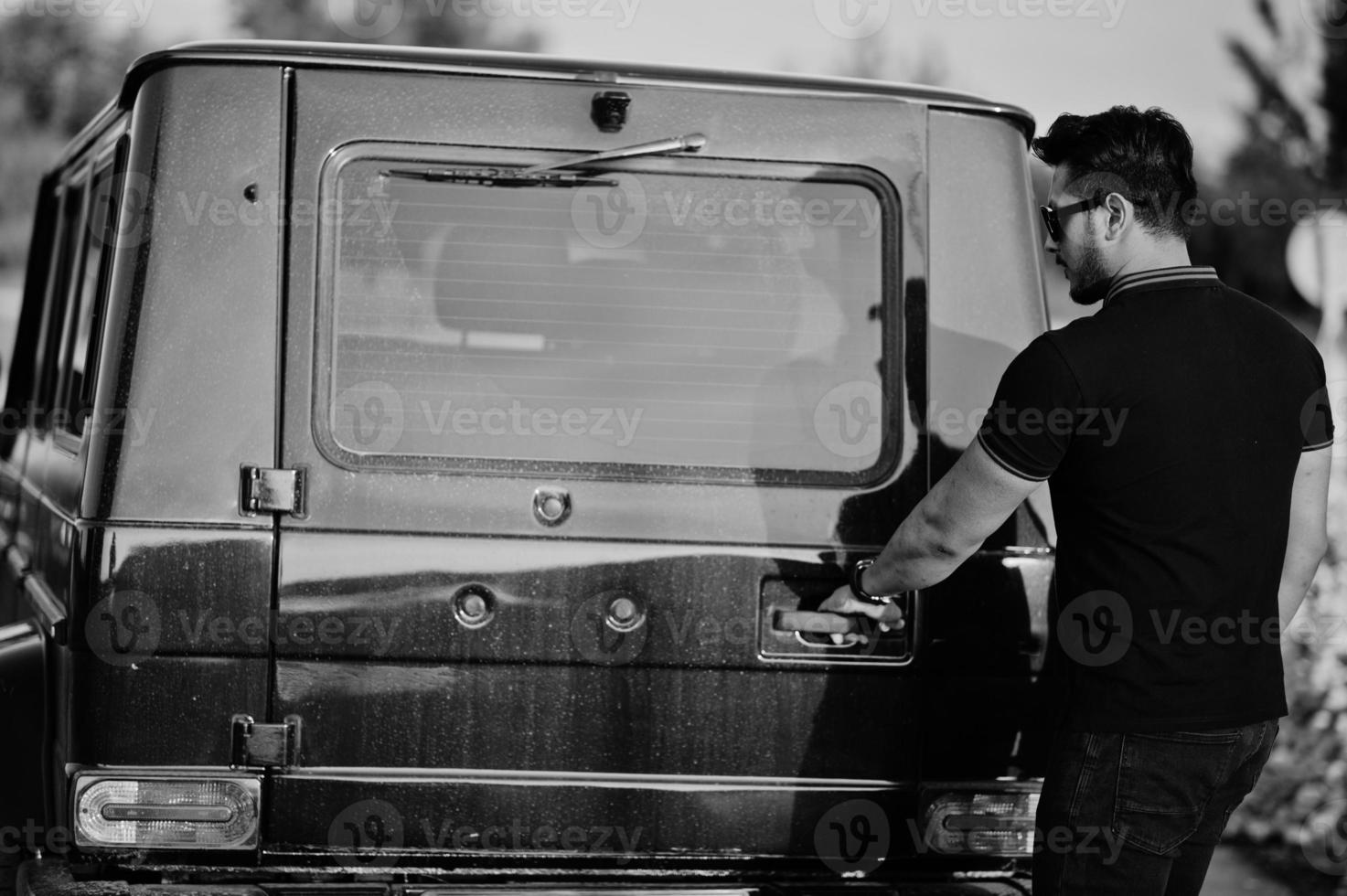 Asian man wear on all black posed near suv car, open rear door. photo