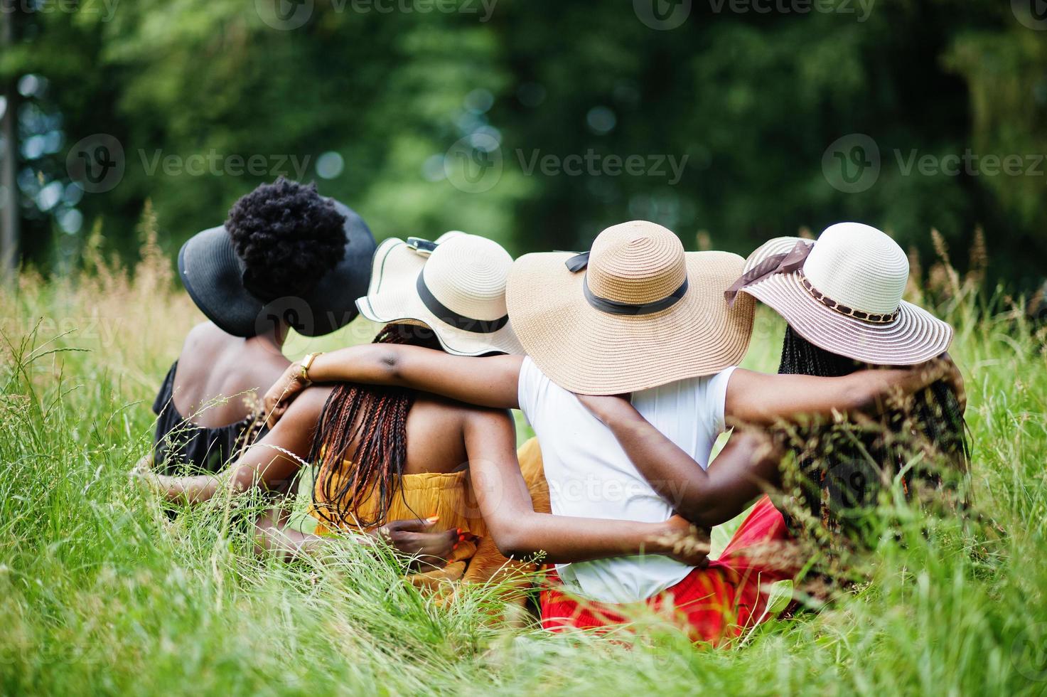 vista posterior del grupo cuatro hermosas mujeres afroamericanas usan sombrero de verano sentadas en la hierba verde en el parque. foto
