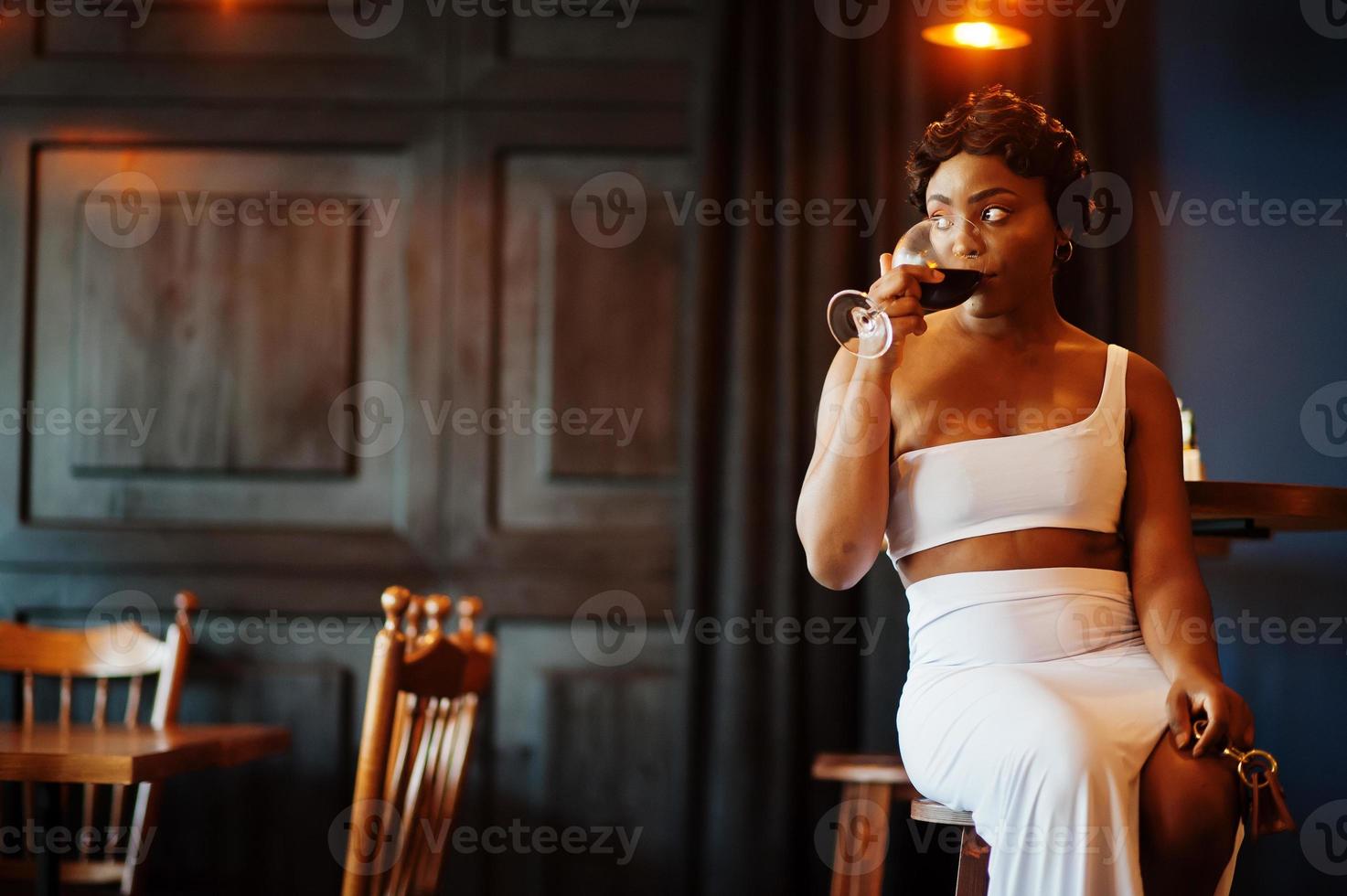 African american woman, retro hairstyle in white dress at restaurant with glass of wine. photo