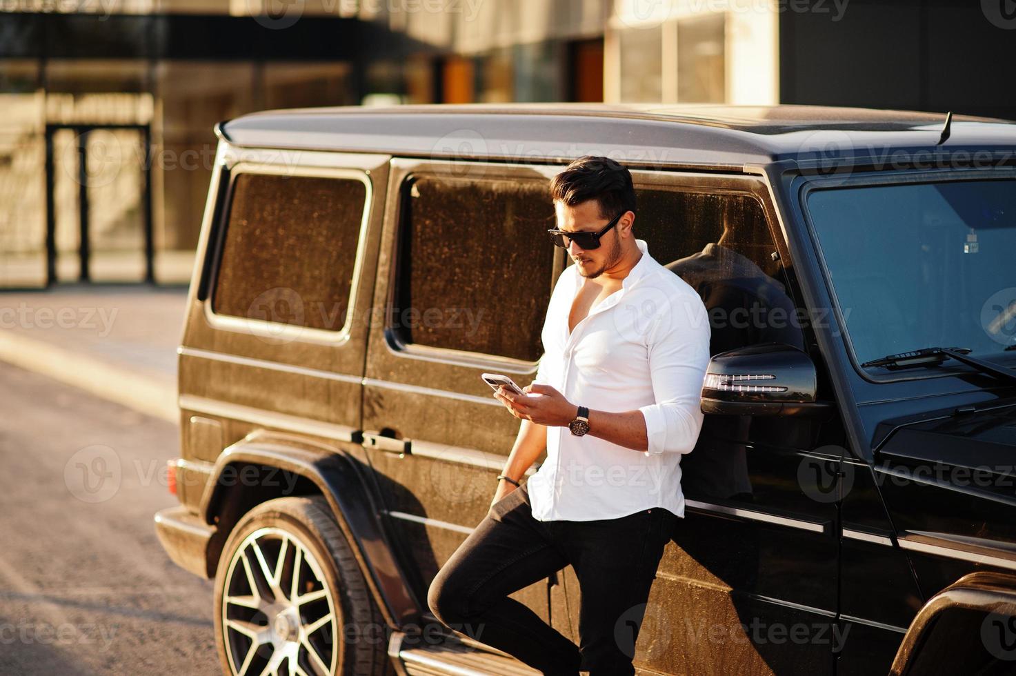 Solid asian man in white shirt and sunglasses posed near black mafia suv car. photo