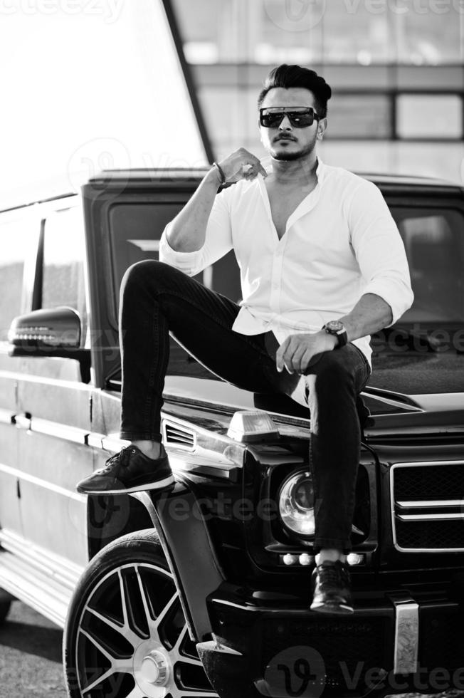 Solid asian man in white shirt and sunglasses posed near black mafia suv car. photo