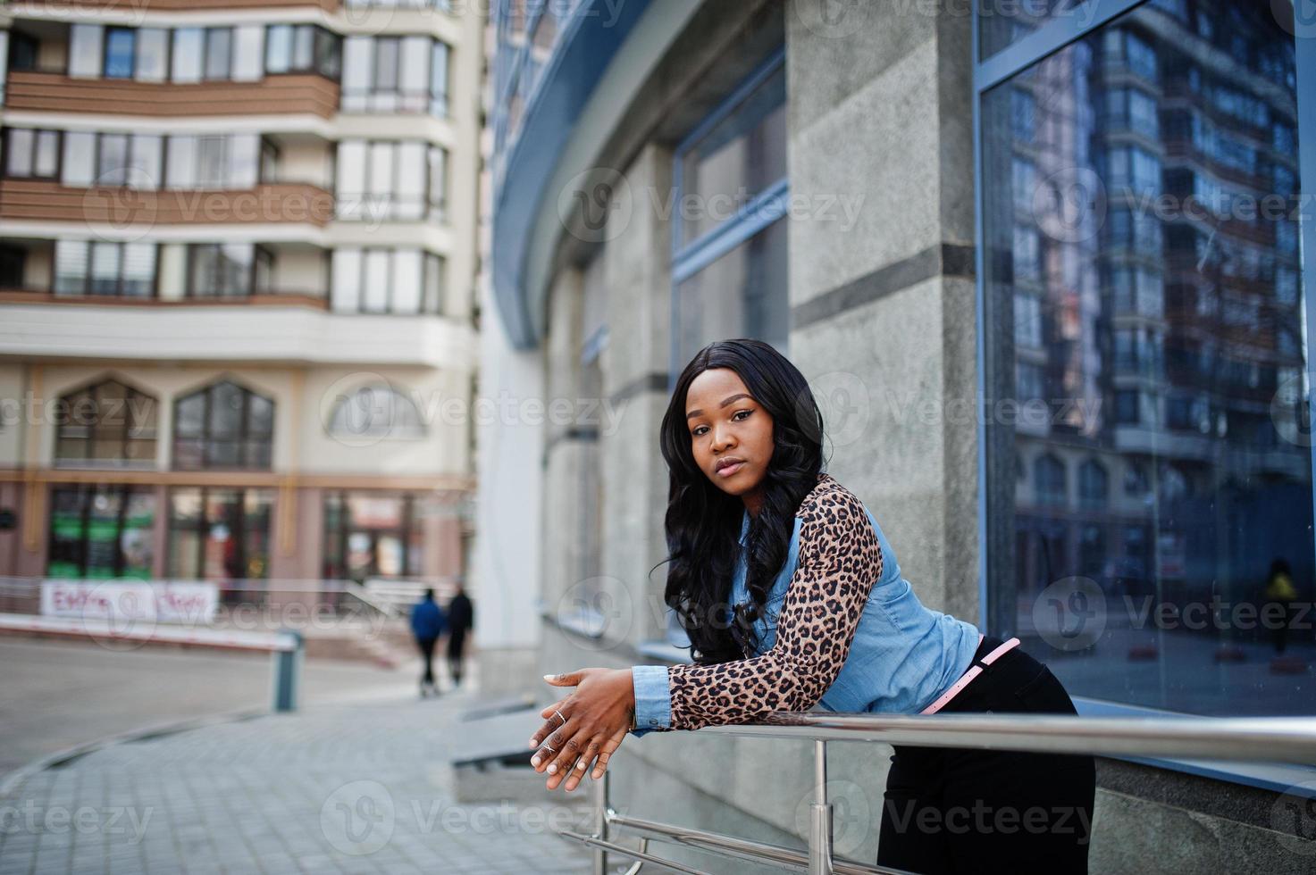 Hipster african american girl wearing jeans shirt with leopard sleeves posing at street against modern office building with blue windows. photo