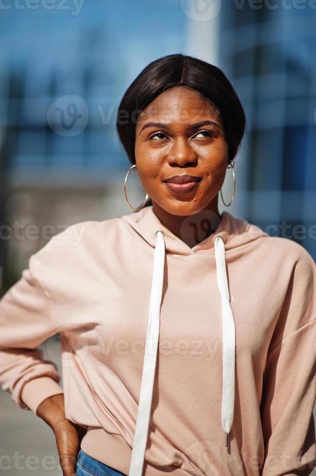 Hipster african american girl wearing pink hoodie, jeans posing at street against office building with blue windows. photo