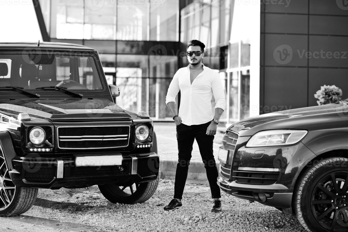 Solid asian man in white shirt and sunglasses posed near black mafia suv cars. photo