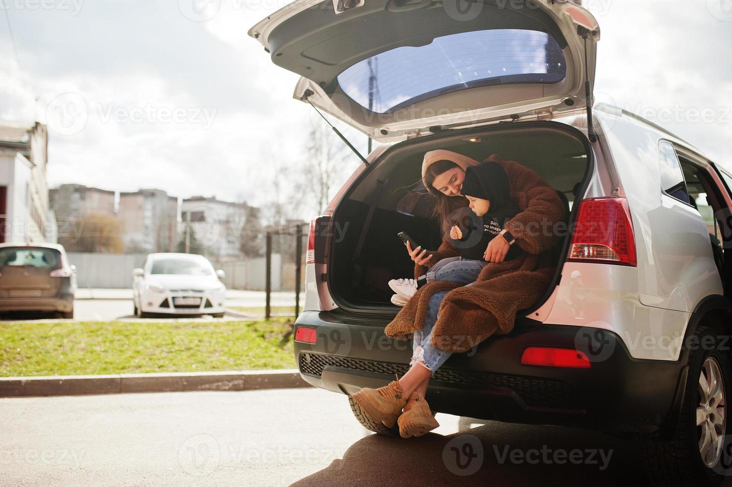 Young mother and child sitting in the trunk of a car and looking at mobile phone. Safety driving concept. photo
