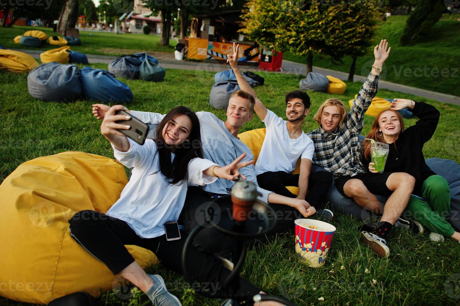 Young multi ethnic group of people watching movie at poof in open air cinema and making selfie on phone. photo
