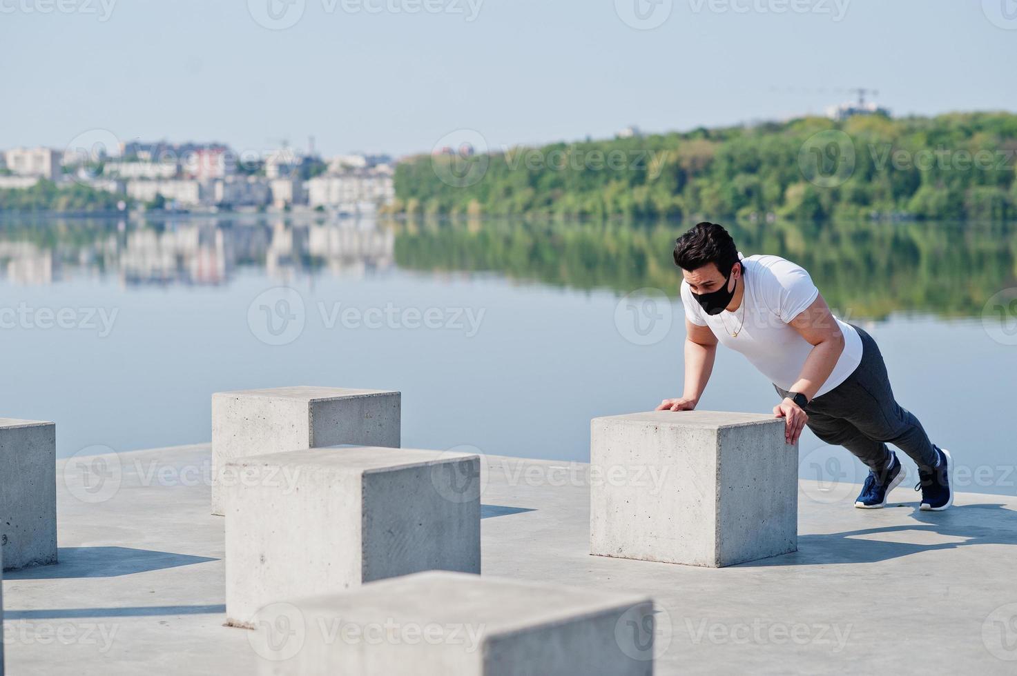 Portrait sports arabian man in black medical face mask doing morning workout exercises against lake during coronavirus quarantine. photo