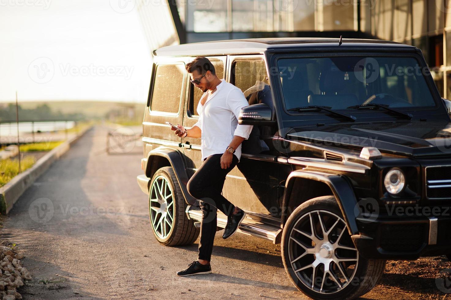 Solid asian man in white shirt and sunglasses posed near black mafia suv car. photo