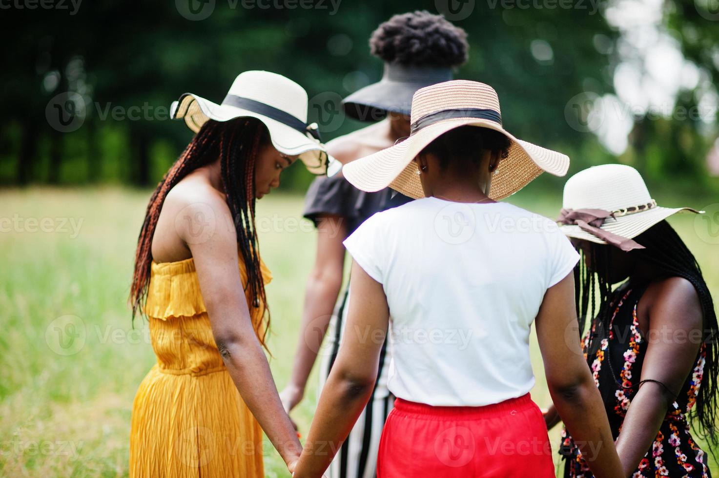 Group of four gorgeous african american womans wear summer hat holding hands and praying at green grass in park. photo