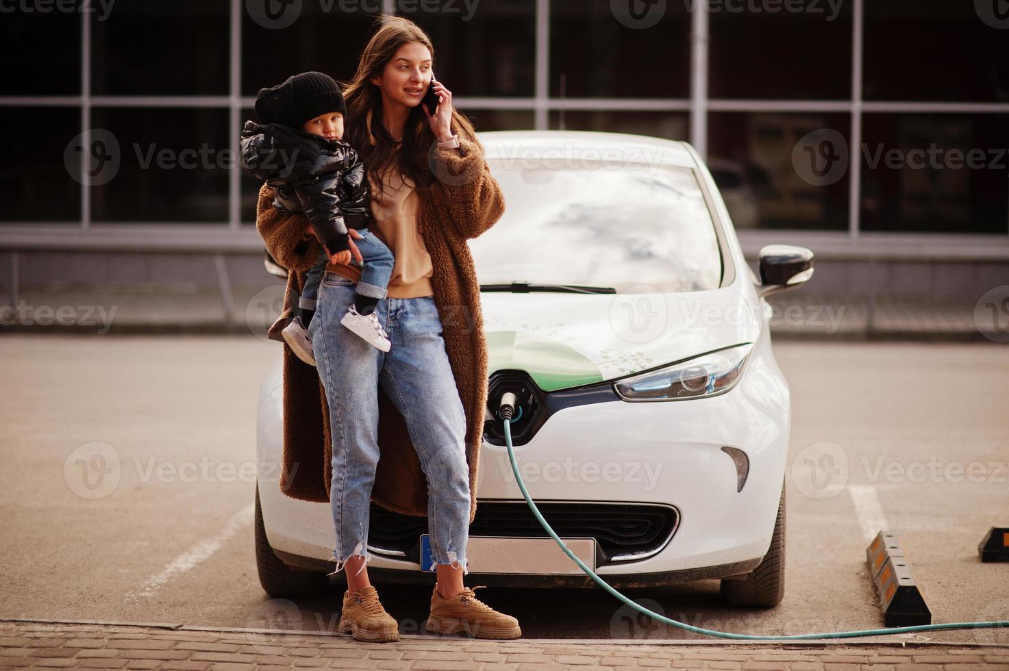 Young mother with child charging electro car at the electric gas station and speak on mobile phone. photo