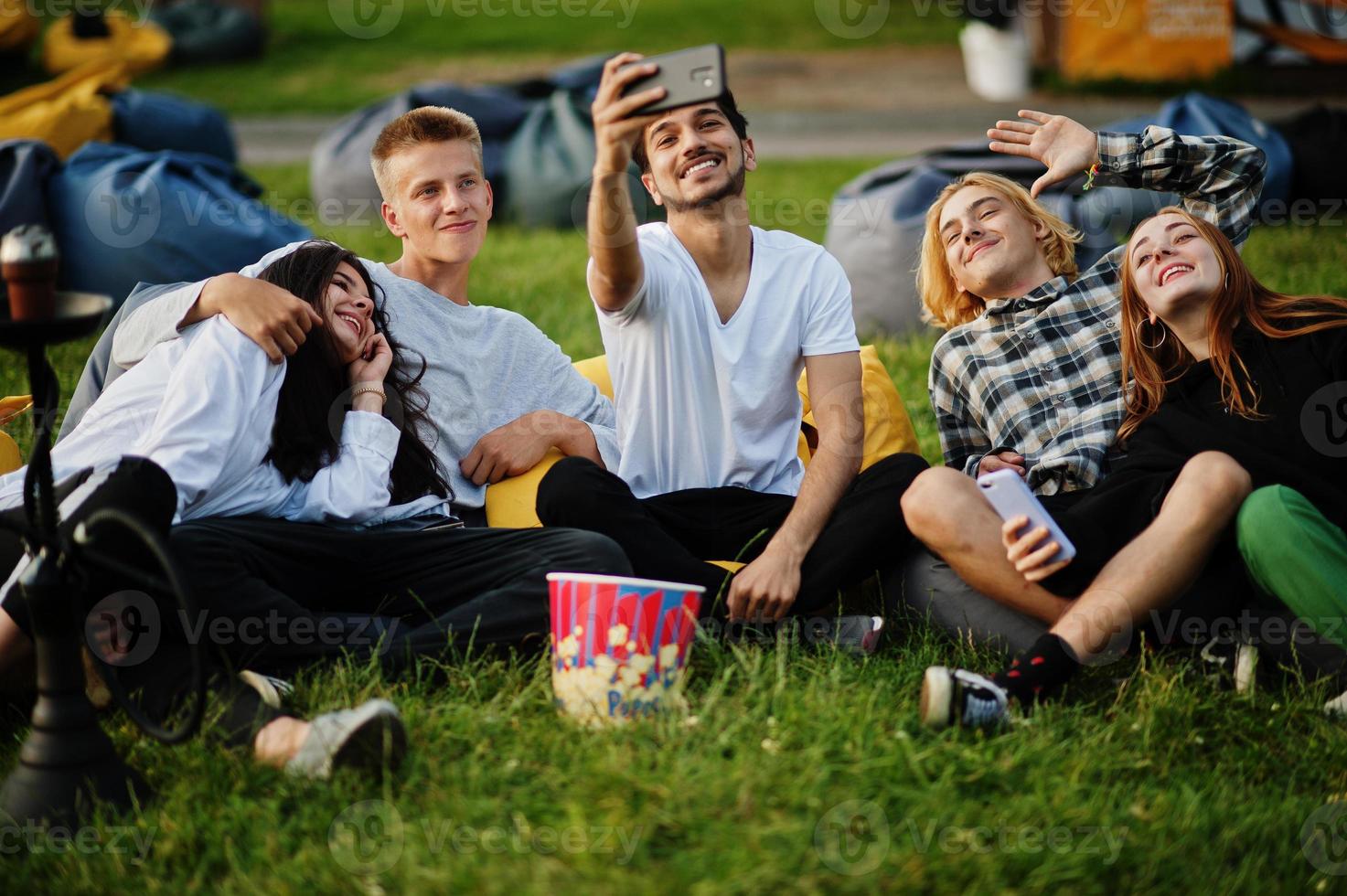 joven grupo multiétnico de personas viendo películas en poof en cine al aire libre y haciendo selfie por teléfono. foto