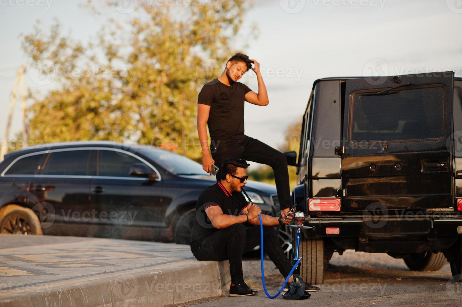 Two asian brothers man wear on all black posed near suv cars and smoke hookah. photo