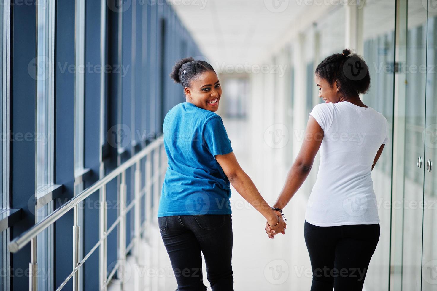 Back of two african woman friends in t-shirts walking halding hands indoor together. photo