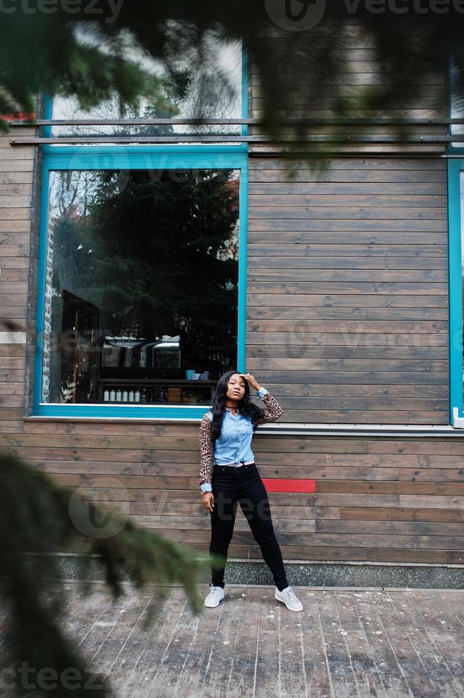 Hipster african american girl wearing jeans shirt with leopard sleeves posing at street against wooden house with windows. photo